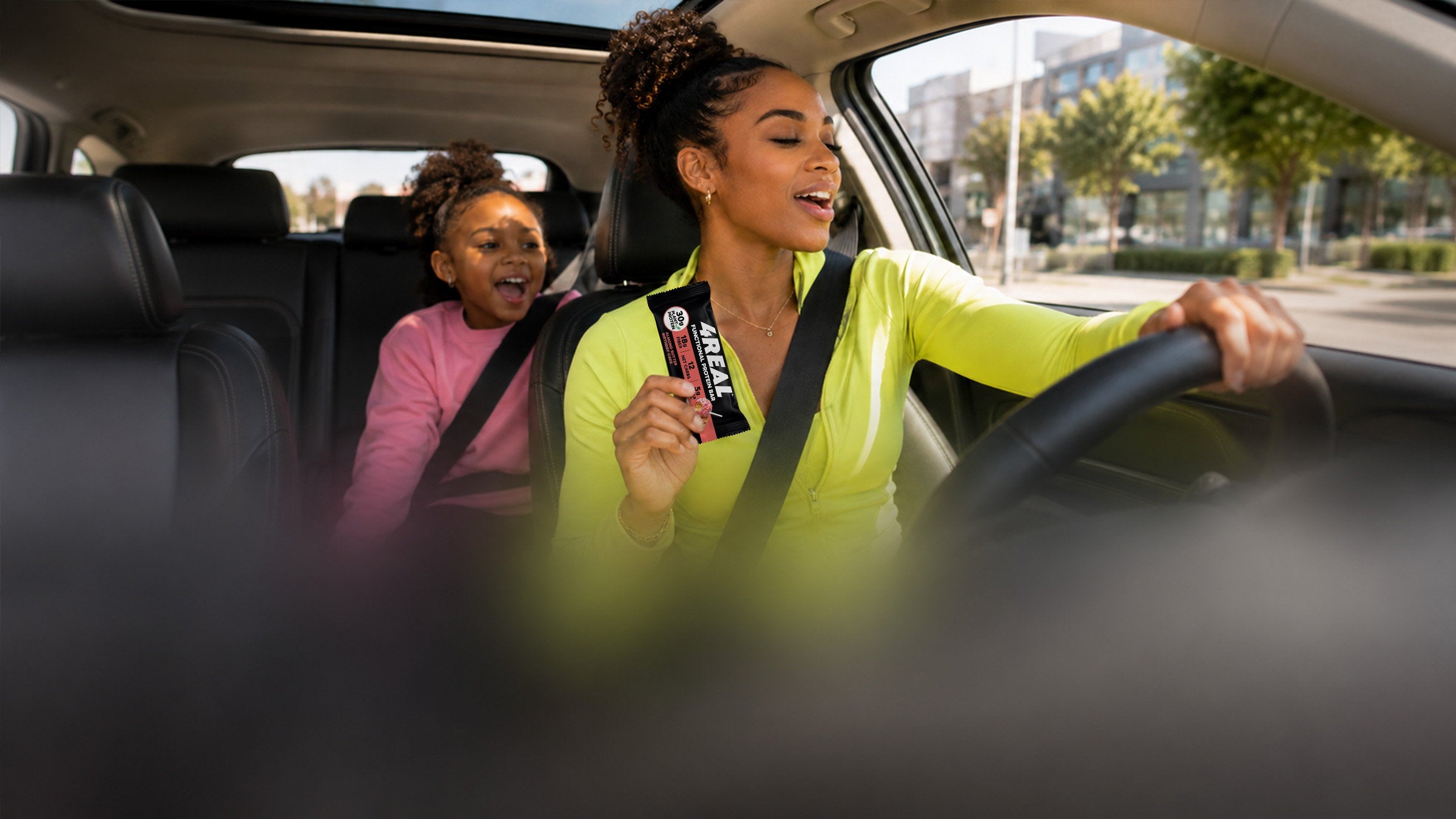A woman singing behind the wheel of a car holds up a 4REAL Almond Butter Birthday Cake bar while a young girl sings along from the back seat, a sunlit city street beyond the windows