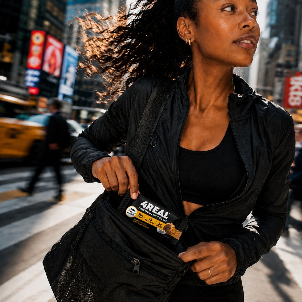 A woman in a black jacket crossing a busy city street pulls a 4REAL Peanut Butter Chocolate Chunk bar out of her black gym duffel, yellow cabs blurred behind her