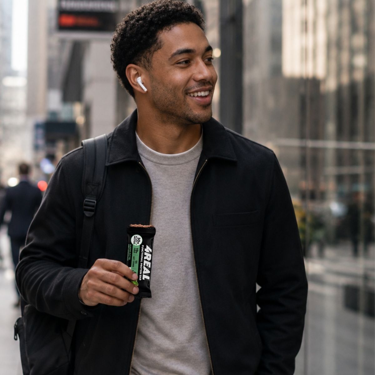 A man in a black jacket and wireless earbuds walks a downtown sidewalk holding a half-unwrapped 4REAL Almond Butter Mint Chocolate bar